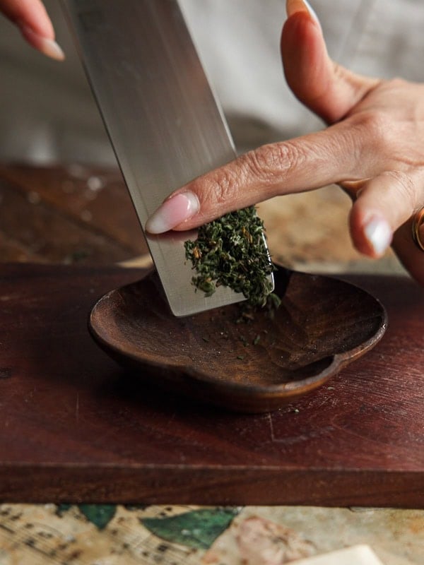 Easy Vegan Lemon Loaf Recipe - Better Than Starbucks 4 A person uses a knife to slide finely chopped herbs from a cutting board into a small wooden dish, guiding the herbs with their finger—perhaps to sprinkle over a freshly baked vegan lemon loaf.
