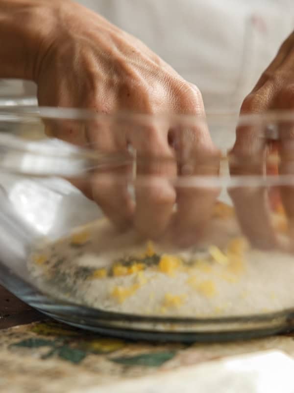Easy Vegan Lemon Loaf Recipe - Better Than Starbucks 9 Close-up of hands squeezing a lemon over a glass bowl filled with cream cheese, eggs, and breadcrumbs, getting ingredients ready to bake a vegan lemon loaf.