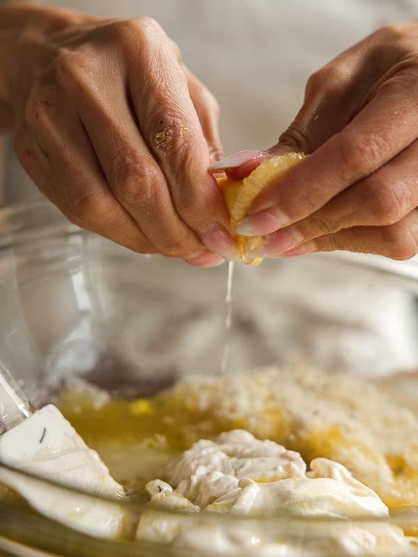 Easy Vegan Lemon Loaf Recipe - Better Than Starbucks 12 Close-up of hands squeezing a lemon over a glass bowl filled with cream cheese, eggs, and breadcrumbs, getting ingredients ready to bake a vegan lemon loaf.