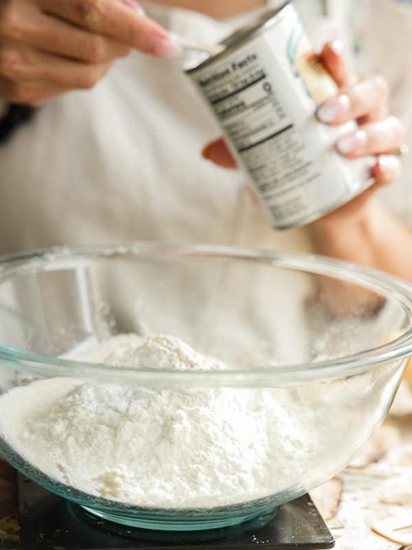 Easy Vegan Lemon Loaf Recipe - Better Than Starbucks 13 A person sprinkles baking powder from a can into a glass bowl filled with flour, preparing ingredients for a vegan lemon loaf. The focus is on the bowl and powder, with hands visible holding the can.