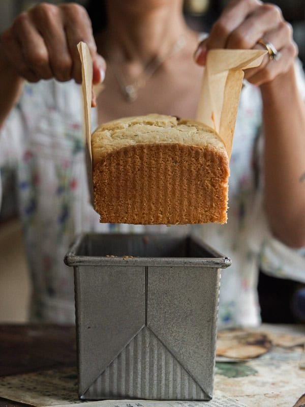 Easy Vegan Lemon Loaf Recipe - Better Than Starbucks 22 A person lifts a freshly baked vegan lemon loaf out of a metal baking pan using parchment paper. The golden brown, rectangular bread is the focus, along with the person's hands carefully holding the loaf.