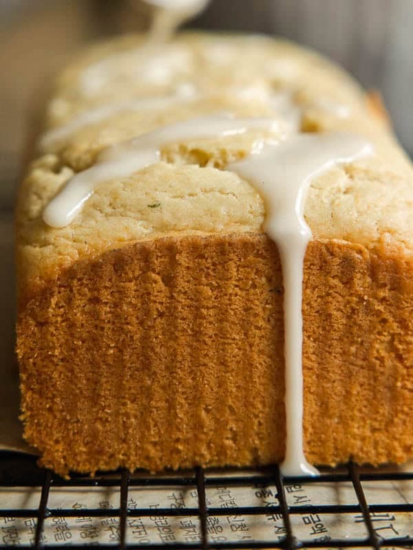 Easy Vegan Lemon Loaf Recipe - Better Than Starbucks 2 A close-up of a golden-brown vegan lemon loaf with white icing drizzled on top, sitting on a cooling rack. The icing is dripping down the side of the loaf.