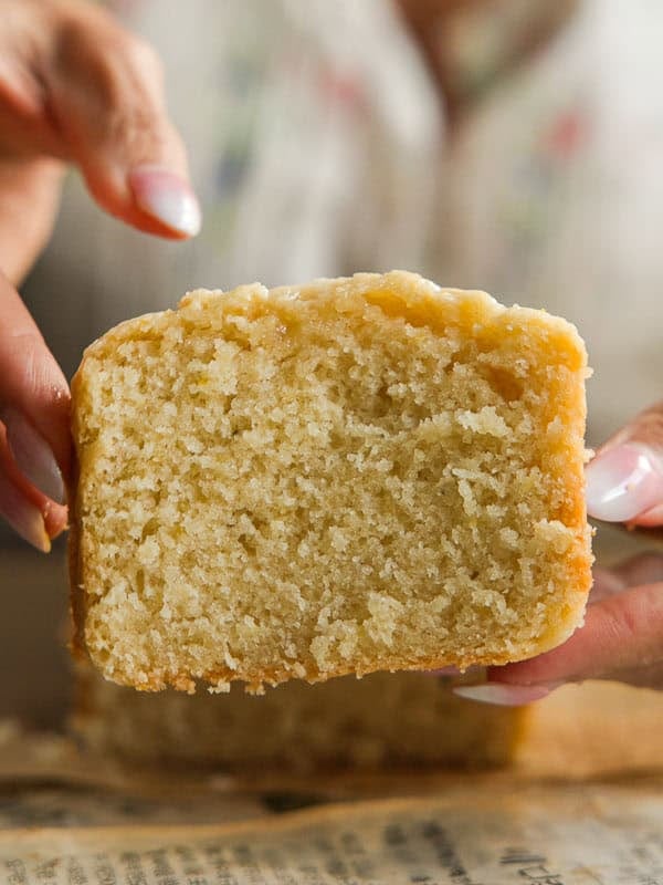 Easy Vegan Lemon Loaf Recipe - Better Than Starbucks 27 A person with light pink nails holds a thick, moist slice of vegan lemon loaf, showing its soft texture and crumb. The background is blurred, focusing all attention on the cake slice.