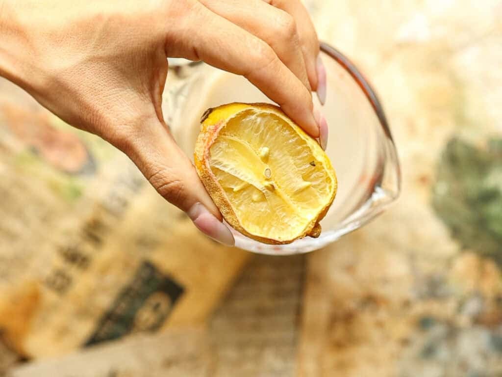 How to Make Viral Salt Bread Recipe (Easy and Vegan). 6 A hand holds a squeezed lemon half over a glass measuring cup, preparing ingredients for an Easy Salt Bread Recipe, with a neutral, blurred background.