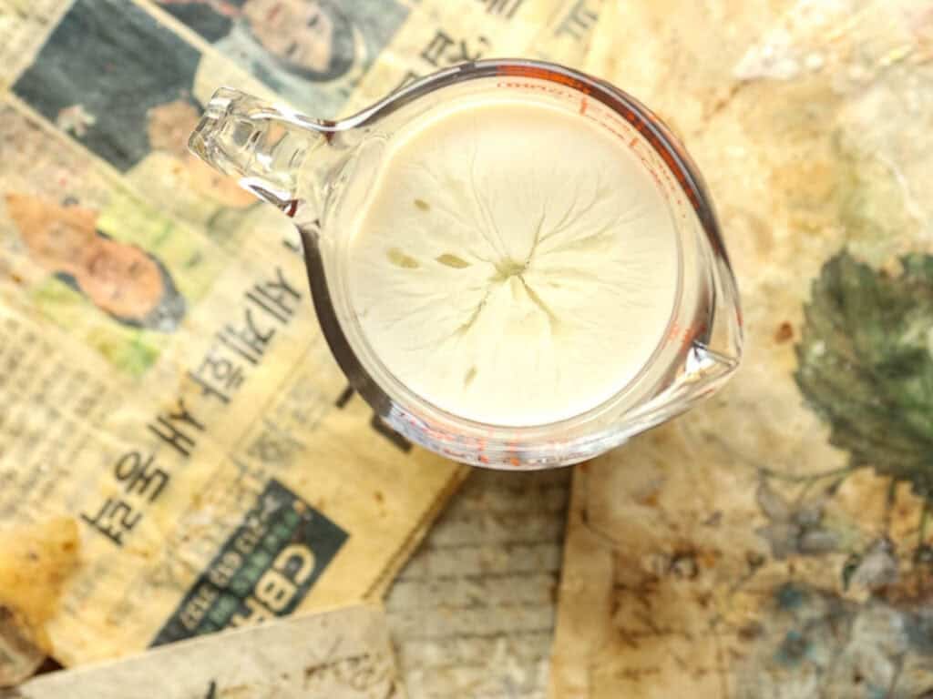 How to Make Viral Salt Bread Recipe (Easy and Vegan). 7 A glass measuring cup filled with a creamy liquid sits on old, yellowed newspaper pages with Korean text and faded images—perfect for preparing your favorite Viral Salt Bread recipe.