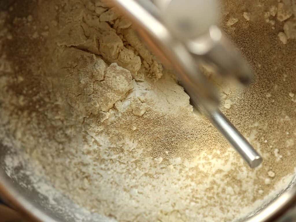 How to Make Viral Salt Bread Recipe (Easy and Vegan). 9 Close-up of flour, yeast, and other dry ingredients in a metal mixing bowl, with a stand mixer attachment above—perfect for starting your vegan bread or salt bread recipe.