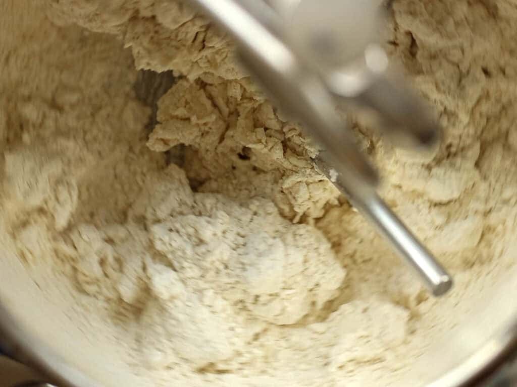 How to Make Viral Salt Bread Recipe (Easy and Vegan). 10 Close-up of dough for a Vegan Salt Bread being mixed in a metal stand mixer bowl, with a paddle attachment blending the ingredients into a crumbly, partially combined texture.