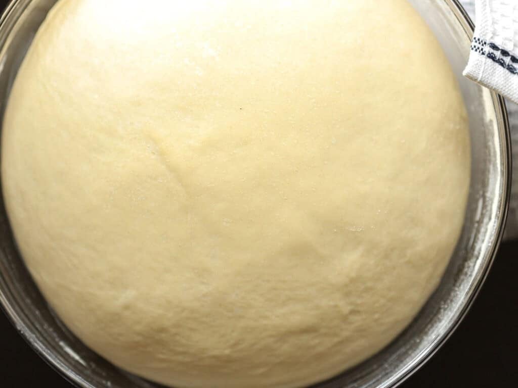 How to Make Viral Salt Bread Recipe (Easy and Vegan). 15 A close-up of risen dough in a glass bowl, perfect for a vegan bread or salt bread recipe. The smooth, pale surface is shown clearly, with a kitchen towel peeking in on the top right edge of the bowl.