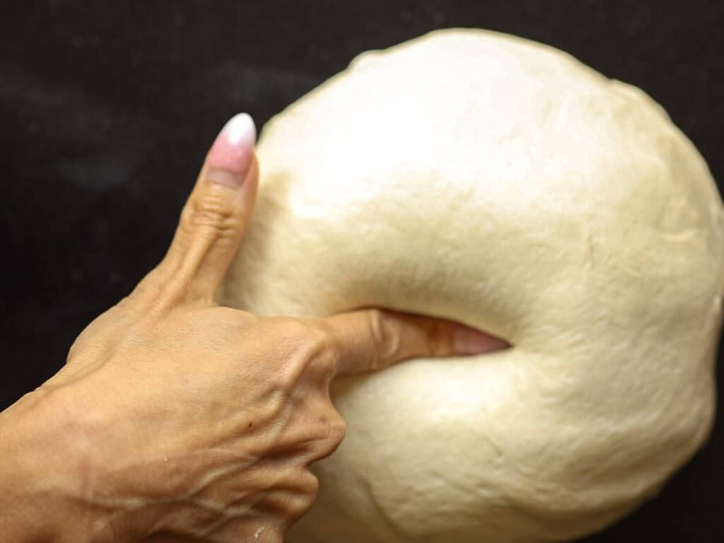How to Make Viral Salt Bread Recipe (Easy and Vegan). 16 A hand presses a finger into a round, smooth ball of dough on a dark surface, testing its readiness for baking this easy salt bread recipe.
