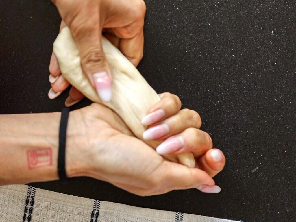 How to Make Viral Salt Bread Recipe (Easy and Vegan). 20 A person with manicured nails kneads dough by hand on a dark countertop, perhaps prepping an easy bread recipe. A black hair tie is on their wrist and a small red tattoo is visible. A striped cloth is partially seen at the bottom of the image.