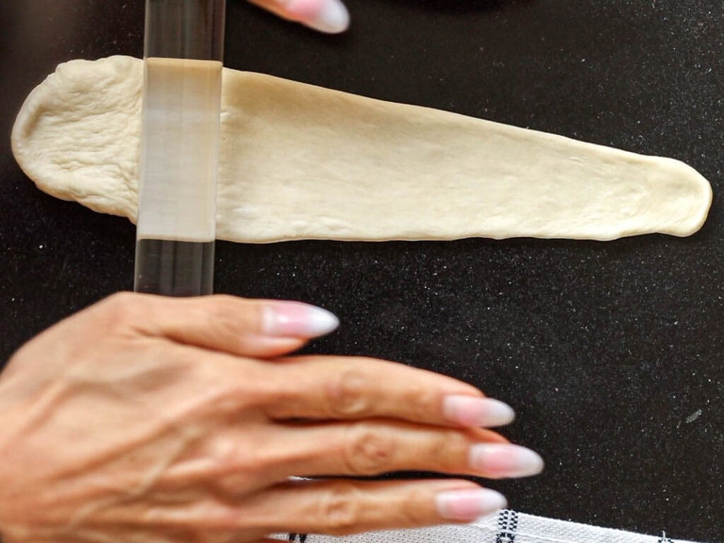How to Make Viral Salt Bread Recipe (Easy and Vegan). 21 A person uses a rolling pin to flatten a strip of dough on a dark surface, preparing a Vegan Bread. Their hands are visible in the foreground, with a striped kitchen towel partially visible at the bottom.