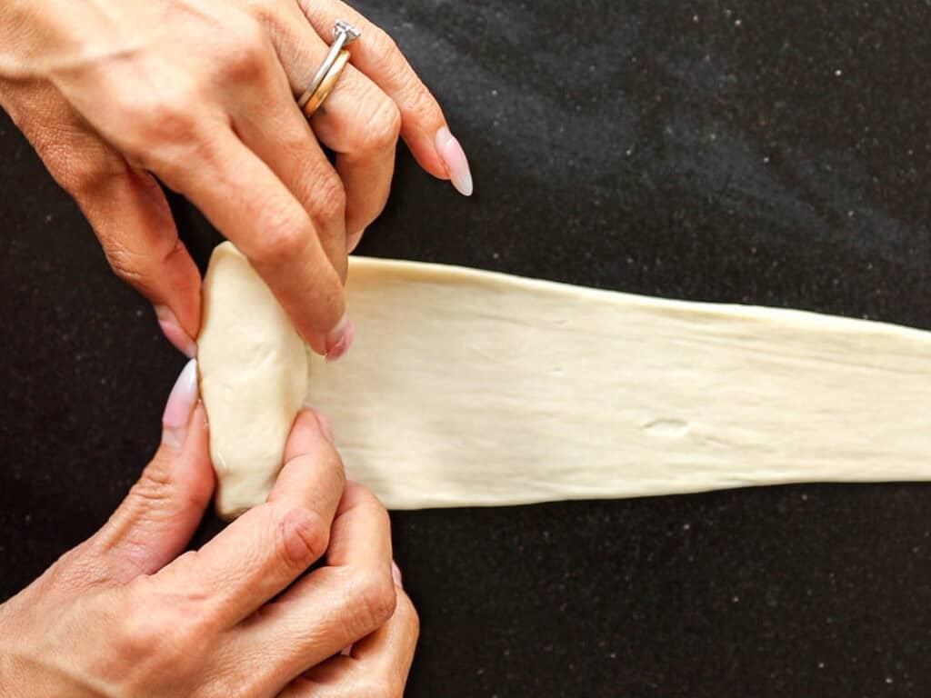 How to Make Viral Salt Bread Recipe (Easy and Vegan). 23 Close-up of hands rolling a strip of dough on a dark surface, preparing it for baking. One hand holds the dough while the other carefully folds the edge inward—perfect technique for making Easy Salt Bread.