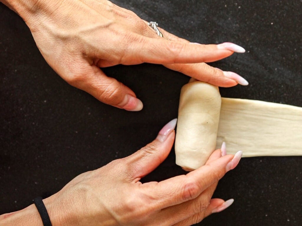 How to Make Viral Salt Bread Recipe (Easy and Vegan). 24 Two hands with manicured nails are rolling a strip of dough to form a compact shape—perhaps crafting salt bread in this viral bread recipe. A ring is visible on one hand and a black hair tie is on the wrist of the other.