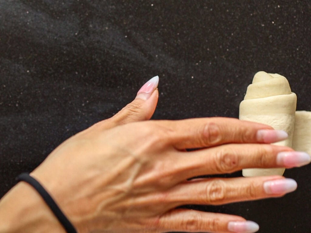 How to Make Viral Salt Bread Recipe (Easy and Vegan). 25 A hand with long, manicured nails rolls viral salt bread dough into a croissant shape on a dark countertop.