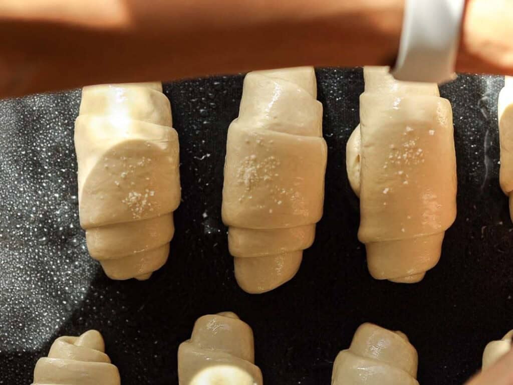 How to Make Viral Salt Bread Recipe (Easy and Vegan). 33 Close-up of several unbaked crescent rolls arranged on a dark baking tray, with a hand holding a brush applying an egg wash to the dough—an easy recipe inspired by viral salt bread.