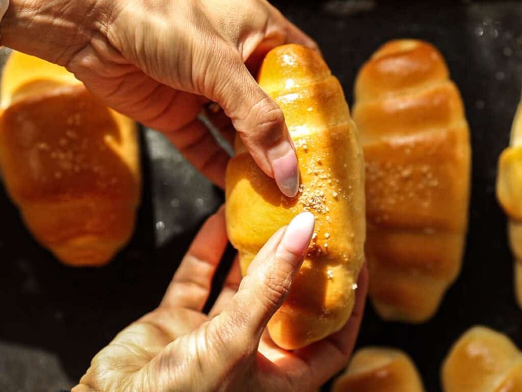 How to Make Viral Salt Bread Recipe (Easy and Vegan). 2 Close-up of hands holding a shiny, golden-brown vegan bread roll, sprinkled with coarse salt, with more similar bread rolls in the background on a dark surface—perfect for anyone seeking an easy bread recipe.