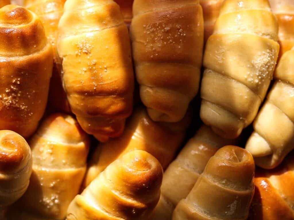 How to Make Viral Salt Bread Recipe (Easy and Vegan). 3 A close-up of several golden brown, shiny, freshly baked bread rolls with a spiral shape, some sprinkled with sugar. These Easy Vegan Bread rolls are tightly packed together and perfect for sharing.