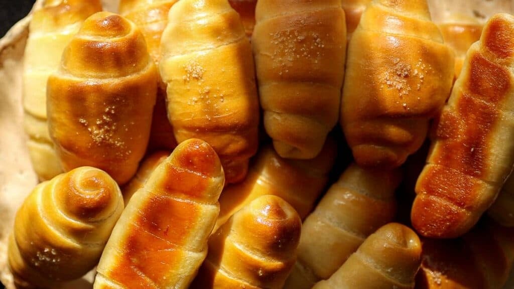 How to Make Viral Salt Bread Recipe (Easy and Vegan). 1 A close-up of several golden-brown, shiny baked rolls—some inspired by the Vegan Salt Bread trend—sprinkled with coarse sugar. The rolls have a smooth, glossy surface and a slightly spiral shape, perfect for trying the viral salt bread recipe.