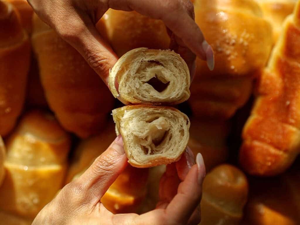 How to Make Viral Salt Bread Recipe (Easy and Vegan). 38 Close-up of hands pulling apart a soft, golden, flaky Salt Bread roll, revealing its airy, layered interior, with more similar pastries blurred in the background.