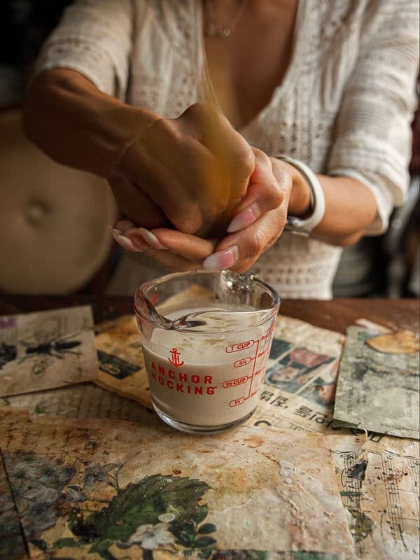 How to Make Viral Salt Bread Recipe (Easy and Vegan). 8 A person squeezes an orange over a glass measuring cup filled with liquid, surrounded by vintage-style papers and a rustic table, perhaps preparing ingredients for a viral bread recipe or homemade vegan bread.