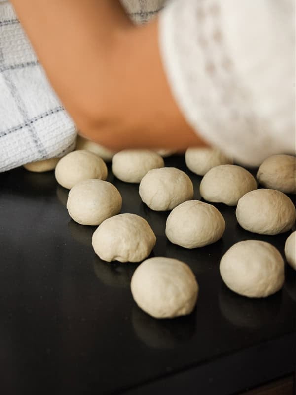 How to Make Viral Salt Bread Recipe (Easy and Vegan). 19 A person’s arm covers a batch of small, round dough balls—perhaps for an easy bread recipe—on a dark countertop with a white kitchen towel. The dough balls are arranged in clusters, ready for the next step in baking.
