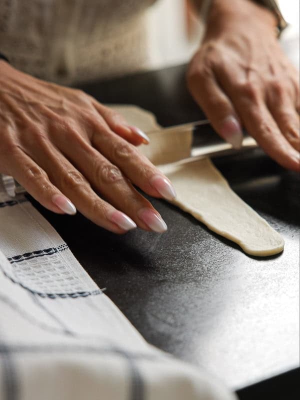 How to Make Viral Salt Bread Recipe (Easy and Vegan). 26 Close-up of hands with manicured nails slicing rolled dough for a vegan salt bread on a dark surface, next to a white kitchen towel with a blue checkered pattern.