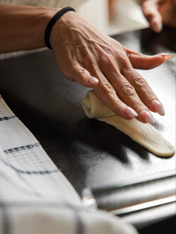 How to Make Viral Salt Bread Recipe (Easy and Vegan). 27 A close-up of a hand rolling dough for an easy salt bread on a dark surface, with a striped cloth nearby and a black hair tie on the wrist.