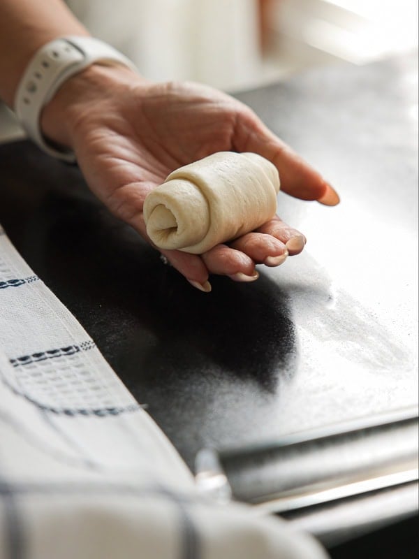 How to Make Viral Salt Bread Recipe (Easy and Vegan). 29 A hand holding a small, rolled piece of dough above a dark countertop, ready to make viral salt bread, with a striped kitchen towel and a rolling pin nearby.