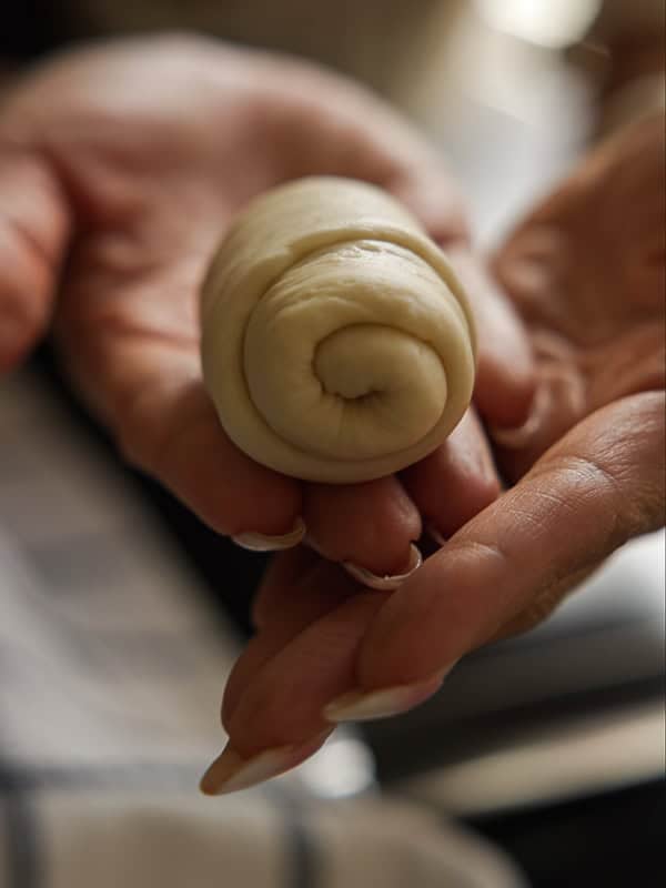 How to Make Viral Salt Bread Recipe (Easy and Vegan). 4 A close-up of hands holding a small, tightly rolled piece of dough with visible layers, ready for baking—perfect for making a Viral Salt Bread. The background is softly blurred.