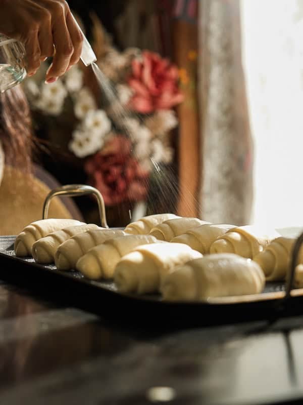 How to Make Viral Salt Bread Recipe (Easy and Vegan). 30 A hand sprays water onto unbaked croissants, arranged on a black baking tray, highlighting steps in a viral bread recipe, with blurred flowers and sunlight softly glowing in the background.