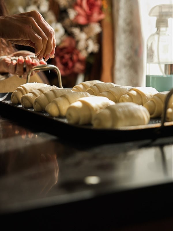 How to Make Viral Salt Bread Recipe (Easy and Vegan). 31 A person sprinkles seasoning on a tray of unbaked, rolled dough pastries while preparing an Easy Salt Bread Recipe in a kitchen, with flowers and a spray bottle visible in the blurred background.