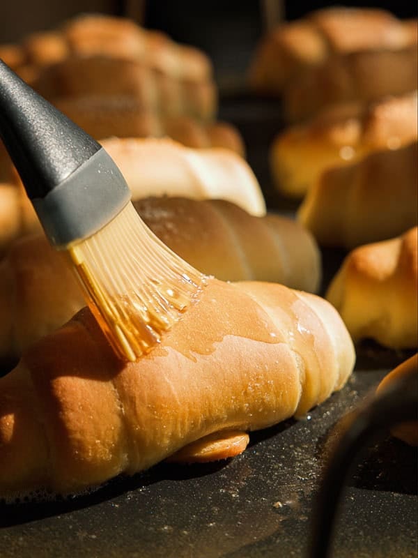 How to Make Viral Salt Bread Recipe (Easy and Vegan). 35 A close-up of a pastry brush applying a shiny glaze to a golden-brown croissant, with several other croissants in the background on a baking tray—perfect inspiration for your next easy bread recipe.