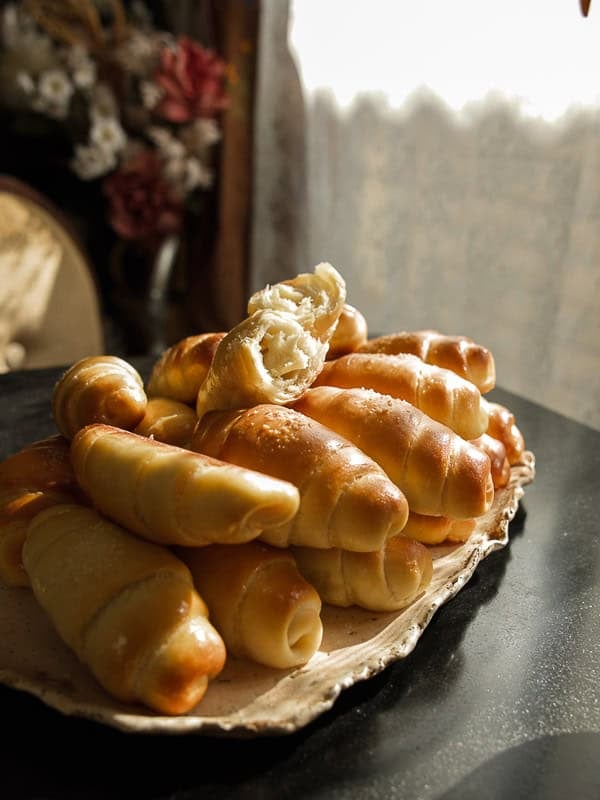 How to Make Viral Salt Bread Recipe (Easy and Vegan). 39 A plate piled with golden, freshly baked bread rolls, one of which is split open to reveal a soft, fluffy interior—perfect for trying your favorite salt bread recipe. Sunlight streams through a lace curtain, warmly illuminating the scene.