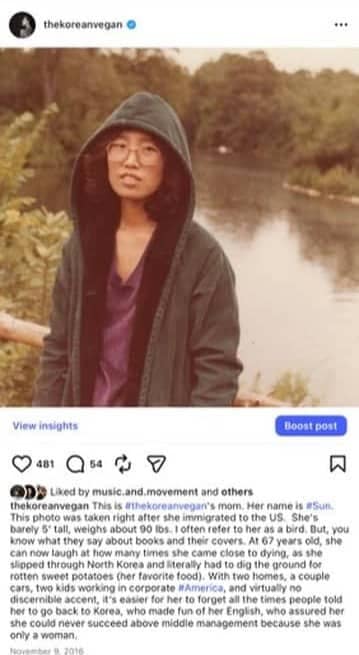 A woman wearing glasses and a hooded jacket stands outdoors near a body of water, with greenery in the background. She faces the camera with a neutral expression, reminiscent of old photographs featuring figures like Barack Obama in candid moments.