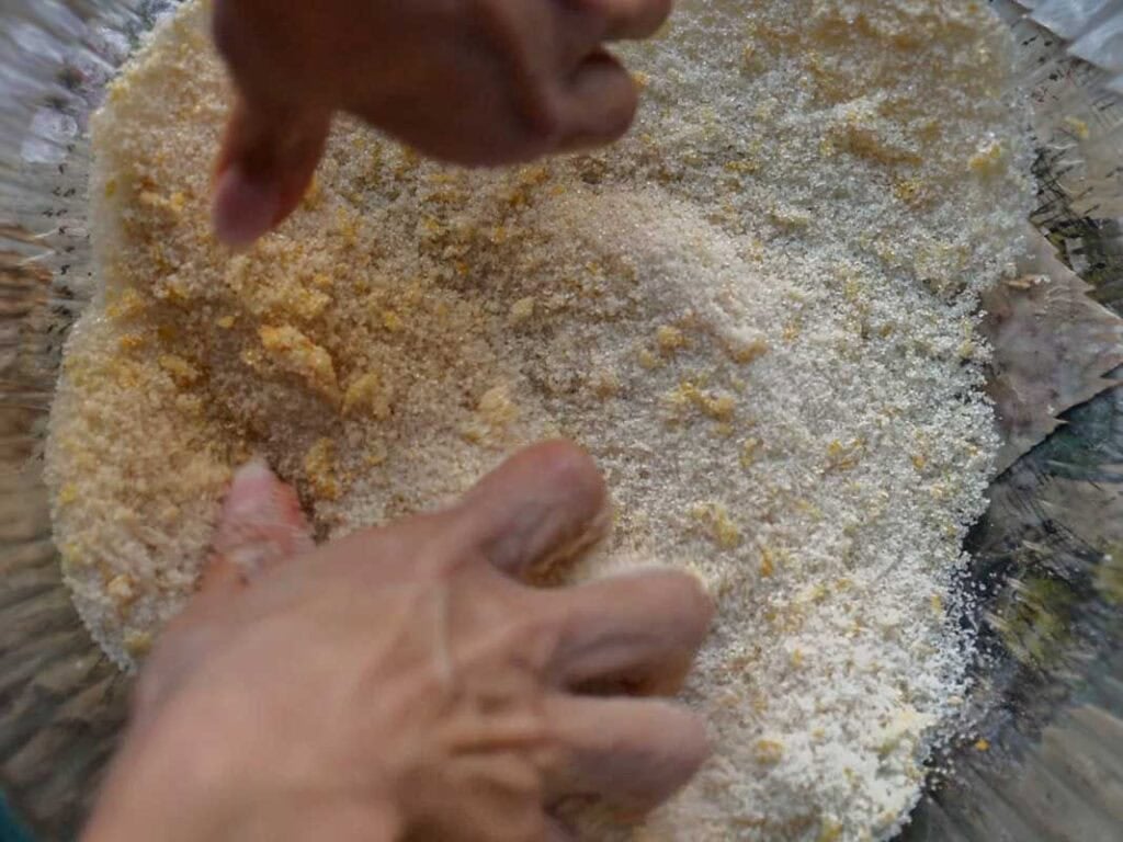 Delicious and Easy Vegan Lemon Poppyseed Muffins. 6 Two hands are mixing sugar and grated ingredients in a large glass bowl, possibly preparing a dough or batter for vegan lemon poppyseed muffins. The mixture appears coarse and slightly yellowish.