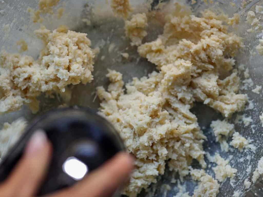 Delicious and Easy Vegan Lemon Poppyseed Muffins. 9 A close-up of cookie dough being mixed in a bowl with a hand holding an electric mixer, partially visible in the lower left corner. The thick, partially blended dough is perfect for baking vegan lemon poppyseed muffins.