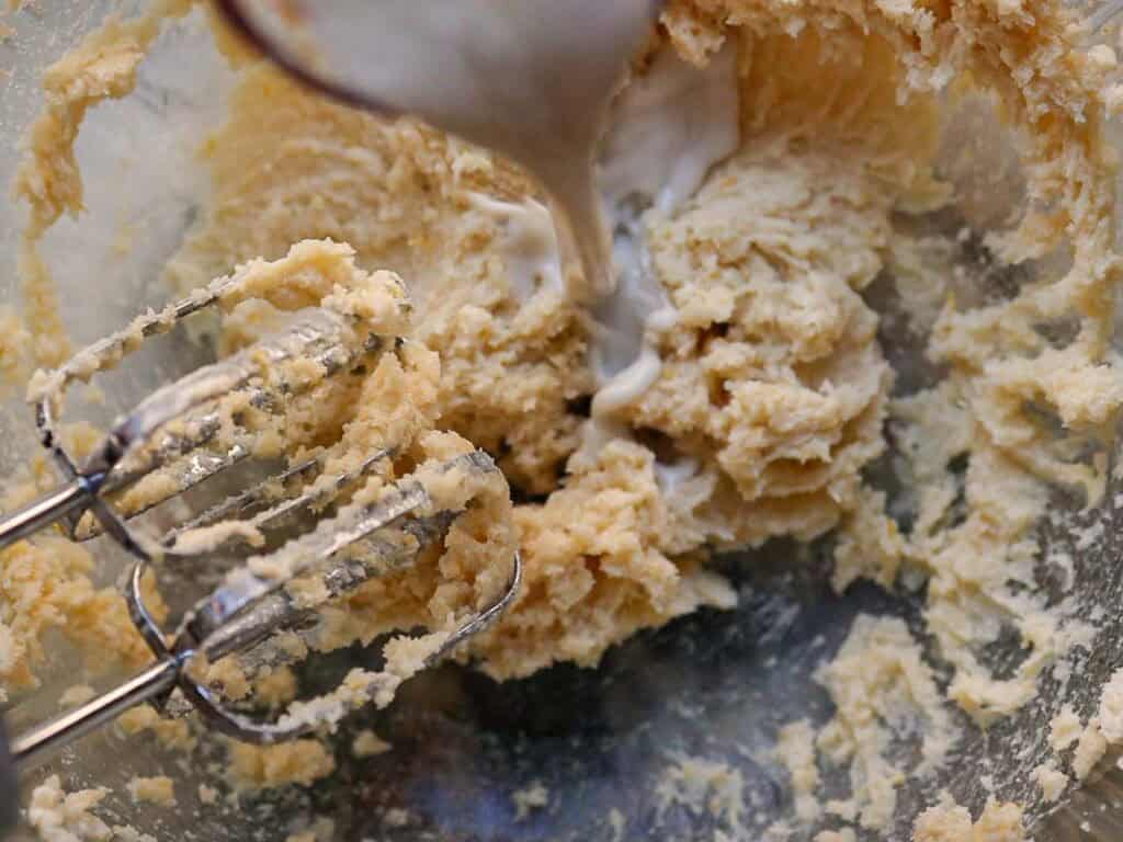 Delicious and Easy Vegan Lemon Poppyseed Muffins. 11 Electric beaters mixing a thick, creamy batter in a glass bowl while liquid is being poured in, likely during baking preparation for vegan lemon poppyseed muffins.