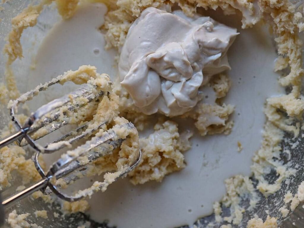 Delicious and Easy Vegan Lemon Poppyseed Muffins. 12 Close-up of a mixing bowl with creamy batter for vegan lemon poppyseed muffins, a dollop of thick cream, and an electric hand mixer with beaters coated in batter.