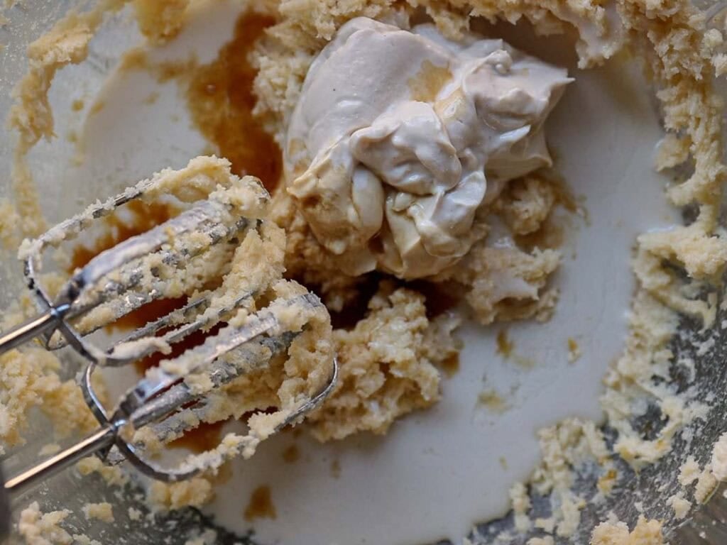 Delicious and Easy Vegan Lemon Poppyseed Muffins. 13 A close-up of an electric mixer with beaters in a bowl containing partially mixed dough and a dollop of cream, perfect for making vegan lemon poppyseed muffins. The mixture appears light in color and creamy in texture.