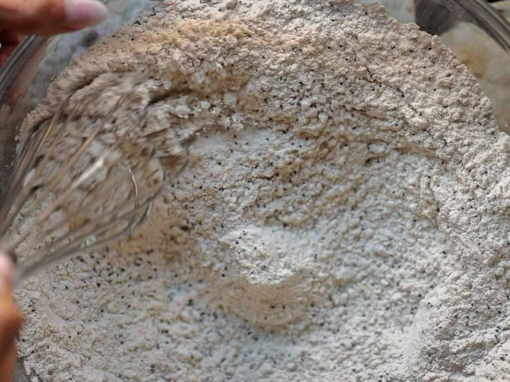 Delicious and Easy Vegan Lemon Poppyseed Muffins. 16 A close-up of hands using a whisk to mix dry baking ingredients, such as flour, in a large glass bowl while preparing vegan lemon poppyseed muffins.