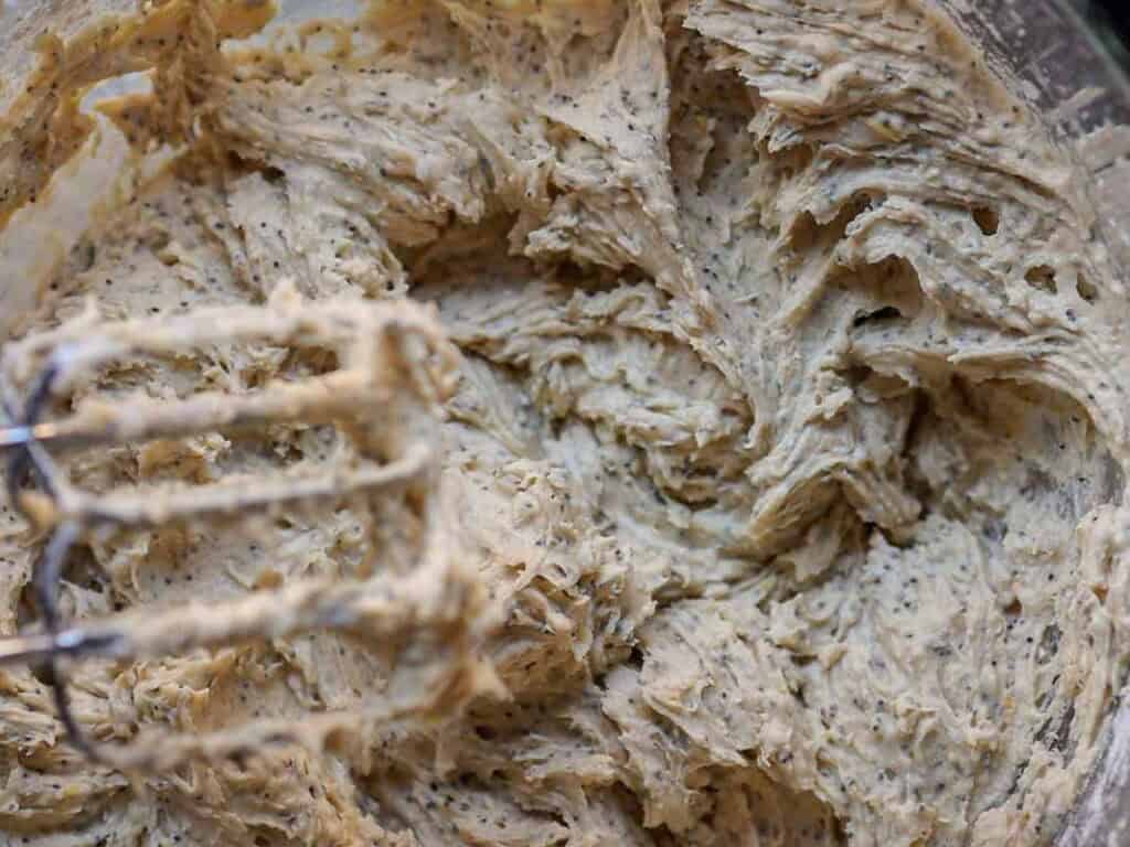 Delicious and Easy Vegan Lemon Poppyseed Muffins. 17 Close-up of creamy, speckled vegan lemon poppyseed muffin batter being mixed in a bowl, with an electric mixer beater visible in the foreground.