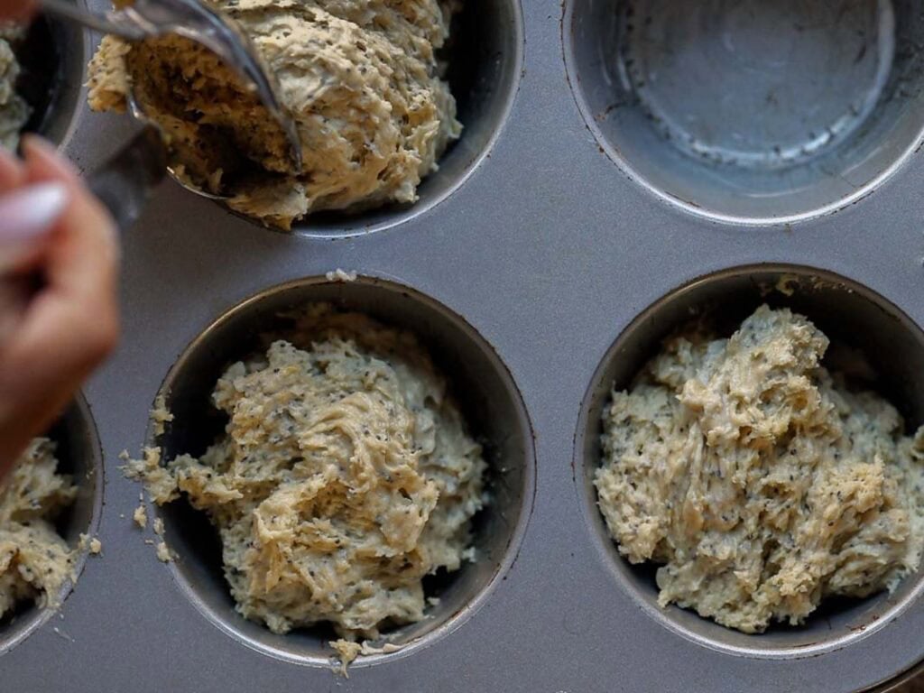 Delicious and Easy Vegan Lemon Poppyseed Muffins. 18 A close-up of a muffin tin being filled with thick, textured vegan lemon poppyseed muffin batter using a scoop. Three cups are filled, and one is empty. A hand holds the scoop over the tin.