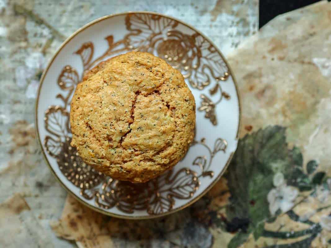 Delicious and Easy Vegan Lemon Poppyseed Muffins. 2 A golden brown, round lemon poppyseed muffin on an ornate white and gold plate, placed on a rustic, patterned surface with earthy tones and floral designsโa scene reminiscent of enjoying freshly baked vegan lemon poppyseed muffins.