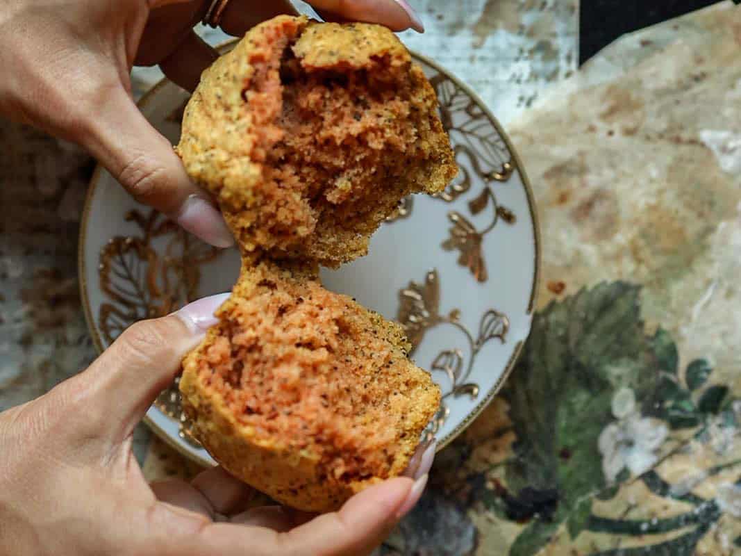 Delicious and Easy Vegan Lemon Poppyseed Muffins. 20 A person breaks open a vegan lemon poppyseed muffin, revealing a moist interior, over a decorative plate with floral patterns on a rustic surface.