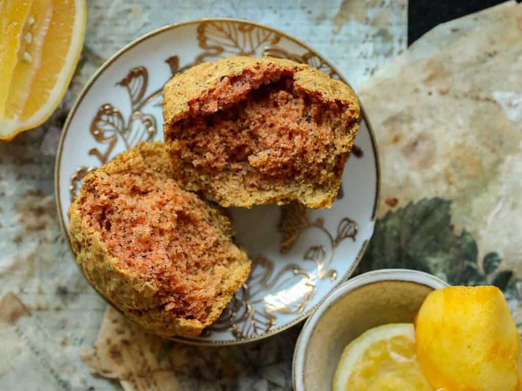Delicious and Easy Vegan Lemon Poppyseed Muffins. 4 A split vegan lemon poppyseed muffin with a reddish-brown interior sits on a decorative plate, surrounded by lemon halves and a cup containing sliced lemon on a textured surface.