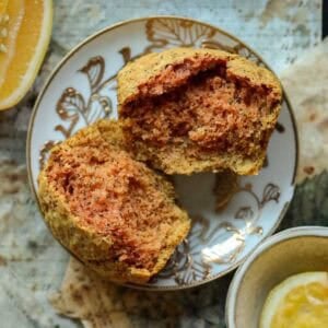 Delicious and Easy Vegan Lemon Poppyseed Muffins. 21 A split vegan lemon poppyseed muffin with a golden-brown, slightly reddish interior sits on a decorative plate, surrounded by slices of citrus fruit on a rustic surface.