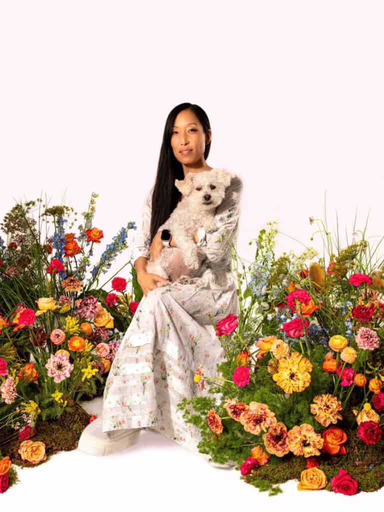 A Short Primer on Birthright Citizenship. 1 A woman in a long, floral dress sits among colorful flowers, holding a small white dog on her lap. The bright background highlights the vivid blooms.