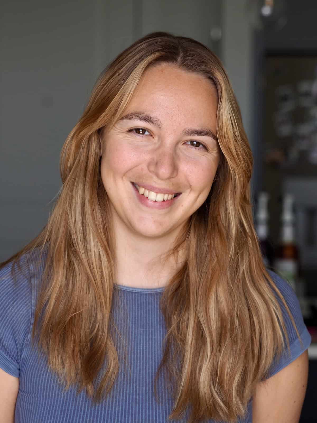 TKV Kollective 3 A young woman with long, light brown hair and a blue ribbed shirt smiles at the camera in a softly lit indoor setting, her Meal Planner open beside her.