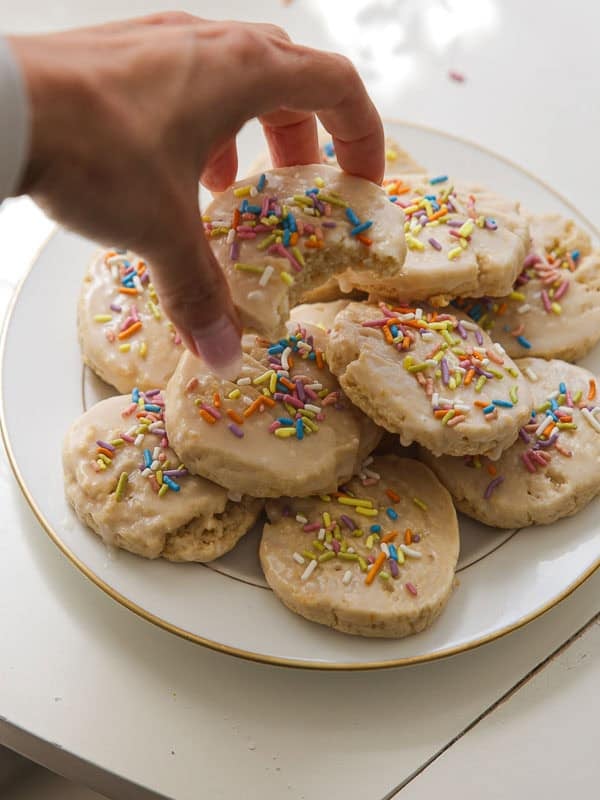 Easy Vegan Lemon Cookies Recipe (Soft and Lemony!) 2 A hand is reaching for a frosted lemon cookie with colorful sprinkles from a plate stacked with similar cookies on a white surface.