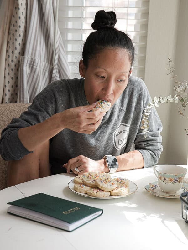 Easy Vegan Lemon Cookies Recipe (Soft and Lemony!) 32 A woman in a gray sweatshirt sits at a table, eating a sprinkle-covered lemon cookie. A plate of lemon cookies, a teacup, and a green notebook are on the table in natural daylight.